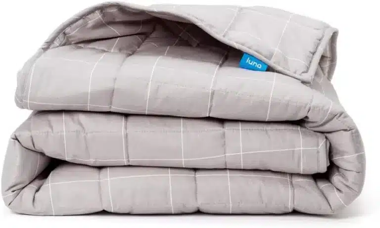 Luna Weighted Blanket Reviews 4 A close-up of a folded light gray Luna weighted blanket, showing the white grid stitching pattern and the blue Luna brand tag. The blanket is softly folded in thirds, creating gentle curves and shadows.